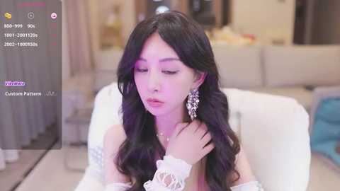 Media: Video of an Asian woman with long, wavy black hair, wearing elegant white lace gloves and dangling earrings, seated indoors with a blurred background.