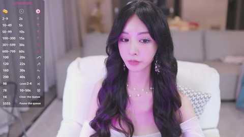 Media: Video of an Asian woman with long, wavy black hair, wearing a white off-shoulder dress, seated in a modern living room with a white couch and grey curtains.