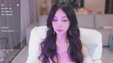 Media: Video of an East Asian woman with long, wavy black hair, wearing a white off-shoulder dress, sitting in a modern living room. The background includes a gray sofa and blurred decor.