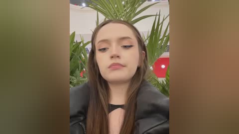 Media: Video of a young Caucasian woman with straight, long brown hair, wearing a black leather jacket, standing in front of lush green plants, with a blurred background.