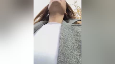 Media: Video of a woman with long, brown hair, wearing a white shirt and a gray cardigan, standing outdoors with blurred background, focusing on her neck and upper chest.