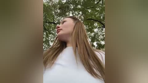 Media: Video of a young woman with long, straight, light brown hair, wearing a white top, standing outdoors against a background of green trees. Her face is serene and turned slightly to the side.