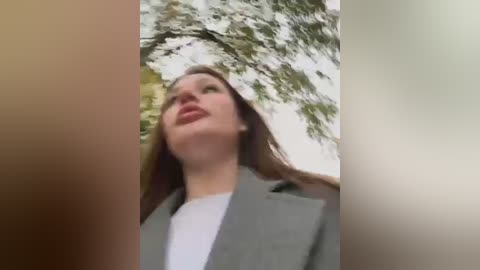 Media: Video of a young Caucasian woman with light brown hair, wearing a gray blazer and white shirt, looking upwards, surrounded by a blurry, leafy tree canopy.