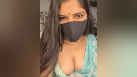 Media: Video of a South Asian woman with long black hair, wearing a black mask, turquoise tie-dye top, and a bindi, in a blurred indoor setting.