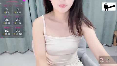 Media: Video of a young Asian woman with straight black hair, wearing a white spaghetti-strap tank top and light skirt, sitting indoors against teal curtains.