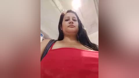 Media: Video of a plus-sized woman with long black hair and a confident expression, wearing a red top, standing in a dimly lit, possibly public restroom.