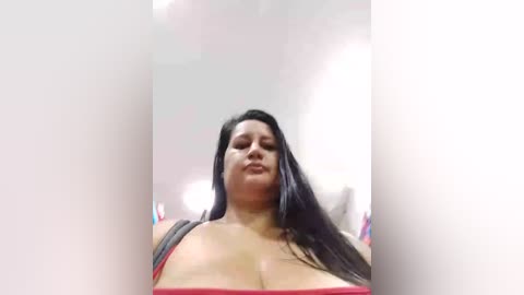 Media: A low-angle video of a plus-sized woman with long black hair, wearing a red strapless top, smiling. The background is a plain white wall with a hint of a light bulb.