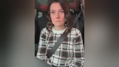 Media: Video of a Caucasian woman with wavy brown hair, wearing a plaid sweater, sitting in a car with a black seatbelt. The background is blurred, showing a dark interior.