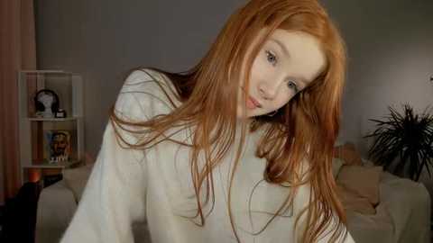 Media: Video of a young, pale-skinned woman with long, red hair and a slender build, wearing a white sweater, leaning forward. Background includes a beige couch, a bookshelf, and a potted plant.