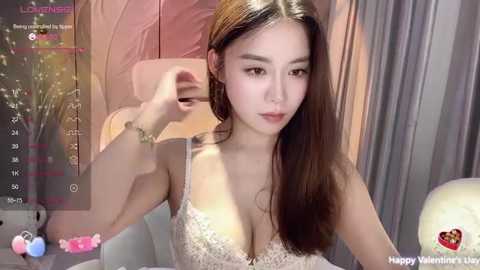 Media: A video of an East Asian woman with long, straight brown hair, wearing a lacy white bra, sitting in a dimly lit room with pink curtains.