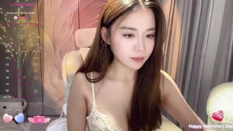 Media: Video of an Asian woman with long, straight brown hair, wearing a delicate, lace camisole, sitting on a bed with pink and beige pillows. Background includes a wall with floral patterns and a lit, spherical lamp.