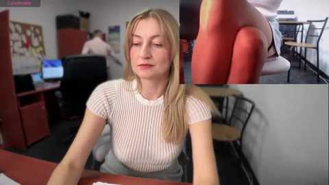 Media: Video of a blonde Caucasian woman in a white ribbed knit top, sitting at a desk, with a close-up of a red chair in the background.