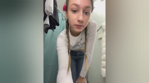 Media: Video of a young Caucasian girl with light skin, brown pigtails, and wearing a white long-sleeve shirt and blue jeans. She leans forward in a bathroom with a teal towel and white towels hanging.