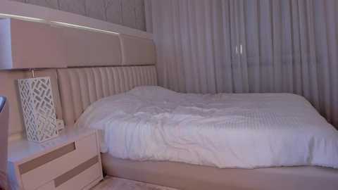 Media: Video of a modern bedroom with a white, quilted bed against a light beige, tufted headboard. A matching white bedside table with a white, geometric-patterned lamp stands next to the bed. The room features sheer, white curtains.