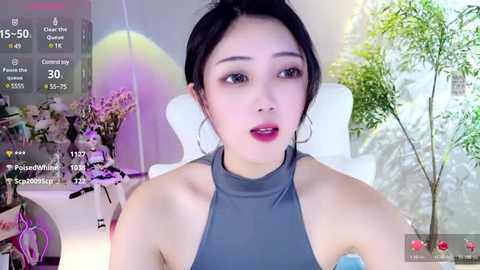 Media: Video of a young Asian woman with fair skin, black hair tied back, wearing a grey halter top, sitting in a modern room with white furniture and green plants, surrounded by virtual chat icons.