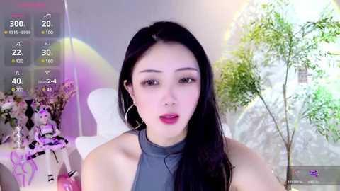 Media: Video of an Asian woman with long black hair, wearing a sleeveless grey top, in a modern room with a white chair and a plant in the background.