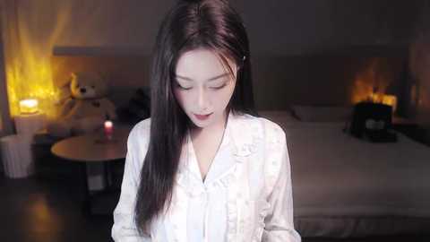 Media: Video of a young Asian woman with long black hair, wearing a white satin robe, standing in a dimly-lit bedroom with a large bed, a lit lamp, and a stuffed teddy bear.