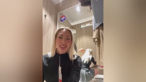 Media: Video of a blonde woman with fair skin smiling, wearing a black jacket, standing in a fitting room with beige walls and mirrors.