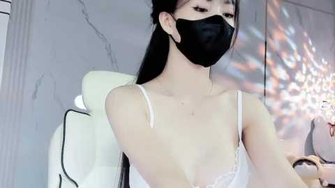 Media: A video of an East Asian woman with long black hair, wearing a white lace camisole and black face mask, seated on a white gaming chair in a modern room with a crystal chandelier.