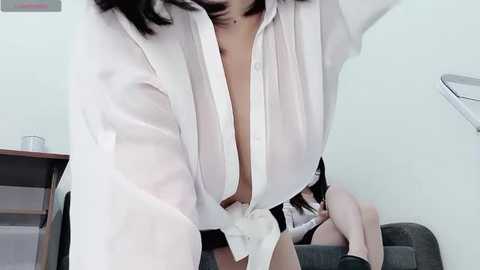 Media: Video of a woman in a white button-up shirt, unbuttoned to reveal black panties, sitting on a gray couch in a minimalist room.