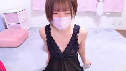 Media: Video of a slender, young Asian woman with straight brown hair, wearing a black lace camisole and a white mask, sitting on a light blue exam table. Background includes pink and white furniture, and a medical cabinet.