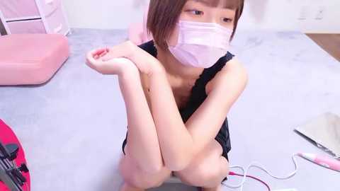 Media: Video of an Asian woman with short brown hair, wearing a surgical mask, black top, and pink scrubs, sitting on a light grey floor, knees drawn up, in a minimalist room with pink furniture and cables.