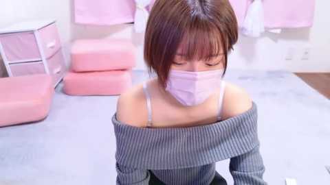 Media: Video of an Asian woman with short brown hair and a mask, wearing a gray off-shoulder sweater, kneeling on a light blue carpet in a room with pink storage bins and pastel-colored items.