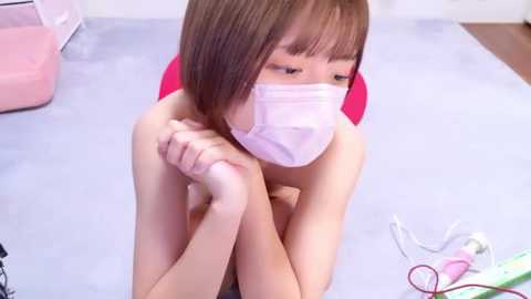 Media: Video of an East Asian woman with short brown hair, wearing a white mask, sitting on the floor with her hands clasped. Background includes a pink chair, a white cable, and a colorful toy.