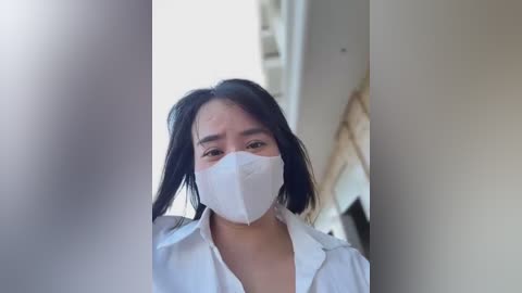 Media: Video of a woman with long black hair, wearing a white mask and lab coat, standing in a bright room with blurred background.