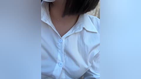 Media: Video of a woman's upper torso in a white button-up shirt, partially obscured by two white objects, likely hands, in a casual indoor setting.