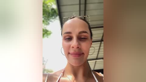 Media: Video of a light-skinned woman with straight brown hair pulled back, wearing hoop earrings, a white halter top, and a smile. Background features a metal canopy and green foliage.