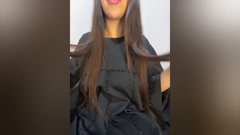 Media: Video of a woman with medium-dark skin tone, wearing a black, glossy, full-length robe with a high collar, her long, straight, dark brown hair partially draped over her shoulders.