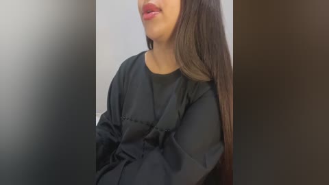 Media: Video of a woman with long, straight brown hair, wearing a black, long-sleeved top, with a slight smile. The background is a blurred, neutral-colored wall.