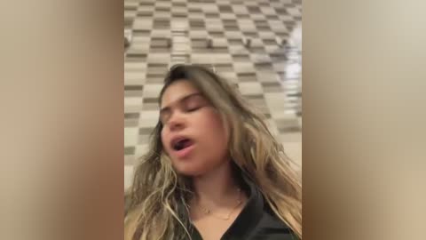 Media: A video of a young woman with long, wavy, blonde hair, wearing a black top, captured in a bathroom with a checkered tile background. She appears to be in a state of ecstasy, mouth open, eyes closed.