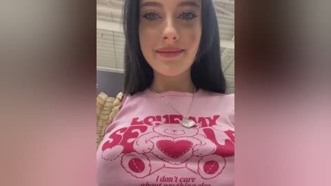 Media: A video of a young woman with long black hair, fair skin, and blue eyes, wearing a pink \"Love Bear\" t-shirt. Background includes a white ceiling and blurry room details.