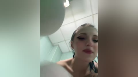 Media: A blurred video of a young woman with wet, dark hair and light skin, standing in a bathroom with white tiles and a ceiling light. She is wearing a white top.