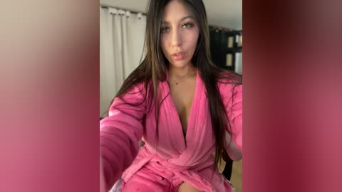 Media: Video of an Asian woman with long black hair, wearing a pink robe, sitting on a bed. Background includes a white curtain and dark furniture.