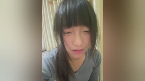 Media: A video of a young Asian woman with long black hair and blunt bangs, wearing a gray shirt, looking down with a neutral expression, indoors with beige curtains and a wooden door in the background.