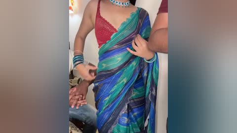 Media: Video of a South Asian woman in a green and blue striped sari with a red lace bra, wearing a blue necklace and bracelets. She stands beside a man with a surprised expression, holding her sari.