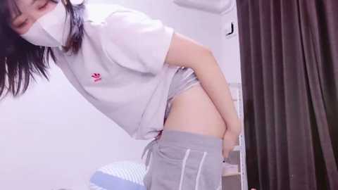 Media: Video of a young Asian woman with light skin and straight black hair, wearing a white face mask and gray sweatpants, bending over in a clinical setting with white walls and medical equipment.