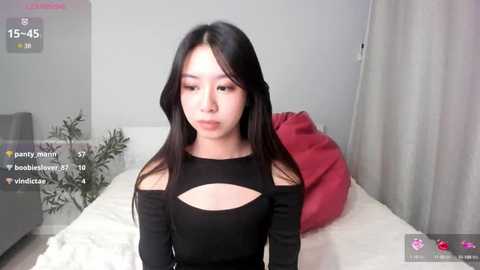 Media: A video of an East Asian woman with long black hair, wearing a black off-shoulder dress, seated on a bed with white sheets, in a modern, minimalistic room.