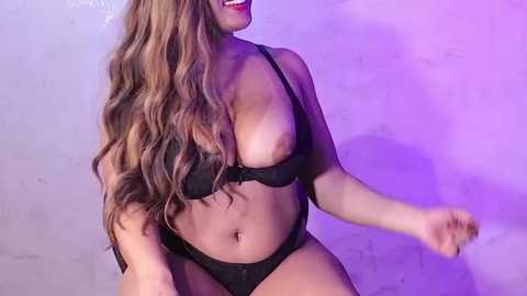 Media: Video of a curvy woman with long, wavy brown hair, wearing a black bra and panties, smiling, against a purple and gray textured wall.