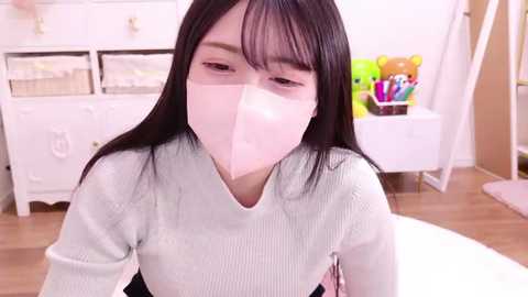 Media: Video of an Asian woman with long black hair, wearing a white surgical mask, light blue sweater, and black pants, in a tidy bedroom with white furniture, stuffed toys, and a round rug.