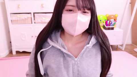 Media: Video of an Asian woman with long black hair, wearing a gray hoodie, pink mask, and white gloves, sitting in a pastel pink room with a white dresser and toy organizer.