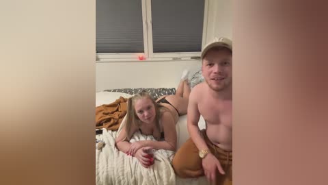 Media: Video of a blonde woman and a man with short hair, both shirtless, lying on a bed with beige sheets. The man has a watch on his wrist.