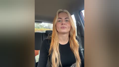 Media: Video of a young woman with long, straight blonde hair, wearing a black top, sitting in the backseat of a car. Outside the window, blurred greenery is visible.