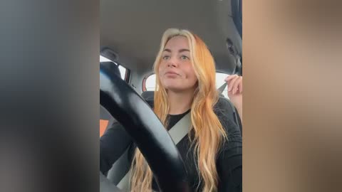 Media: Video of a blonde woman in a black shirt, wearing a seatbelt, seated in a car with gray seats.