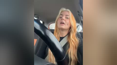 Media: Video of a blonde woman with long hair, wearing a black latex outfit, driving a car, seen from the side. The image is cropped and pixelated, with a blurry background.