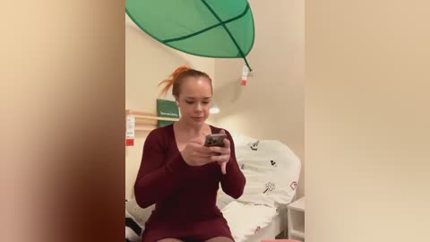 Media: Video of a smiling woman with auburn hair in a ponytail, wearing a maroon sweater, sitting on a white beanbag chair. A large green leaf decoration hangs above her in a beige room with a green wall sign.