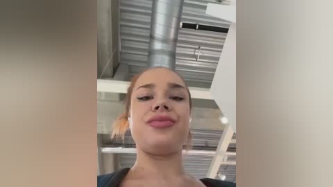 Media: Video of a young woman with fair skin, blonde hair tied back, wearing minimal makeup and a black top. She has a neutral expression and is captured from a low angle in an industrial, metallic, ceiling-lit setting.